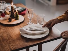 A person sets a white plate with a folded napkin and cutlery on a wooden table decorated with candles and Christmas ornaments, drawing inspiration from catalog resources to create an inviting holiday atmosphere.