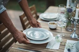 A person sets a patterned plate on top of a larger white plate on a wooden outdoor table with glasses, flowers, and a small bowl, creating an inviting tablescape that could inspire any Catalog Resources collection.