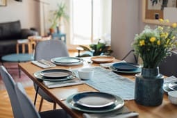 A dining table set for four with plates, bowls, and cutlery, topped with a vase of yellow flowers in a modern, brightly lit living room—an inviting scene perfect for showcasing in Catalog Resources.
