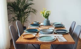 A wooden dining table set with blue and neutral plates, silverware, cloth napkins, and a vase of yellow and white flowers is beautifully arranged for an inviting meal. Six gray chairs surround the table, while a plant in the background adds a touch of greenery. Perfect for showcasing in Catalog Resources, this setting highlights both elegance and comfort.