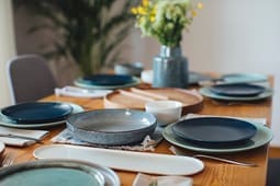 A wooden dining table set with plates, bowls, and cutlery is beautifully arranged, looking ready for a meal. A vase with flowers sits in the center, adding a fresh touch to the inviting setting—perfect for featuring in your Catalog Resources.