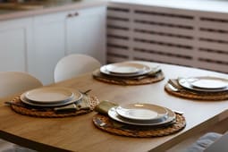 A wooden dining table set for four with plates, bowls, cutlery, and napkins on woven placemats in a modern kitchen, perfect for showcasing in Catalog Resources.
