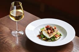 A glass of white wine sits next to a white plate with a piece of fish topped with vegetables, served on greens and a brown wooden table—an inviting scene perfect for showcasing in your Catalog Resources.