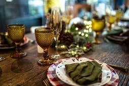 A wooden table set for a meal with patterned plates, green napkins, yellow glasses, champagne flutes, and festive decorations with lights is the perfect inspiration for your next gathering—explore more ideas in our Catalog Resources.