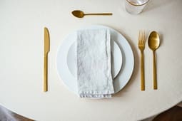 A table setting with a white plate, a folded napkin, gold-colored cutlery, and a glass of water on a light tablecloth—ideal inspiration for Catalog Resources seeking elegant dining arrangements.