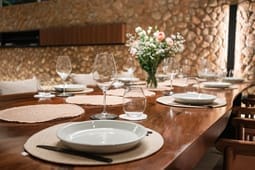 A wooden dining table is set with plates, glasses, and cutlery. A vase of flowers is in the center. The background features a stone wall, creating a scene reminiscent of those found in catalog resources.