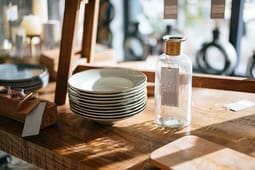 Stack of ceramic plates and a glass bottle with a gold cap on a wooden table in a sunlit shop or kitchen setting, perfect for showcasing in your Catalog Resources.