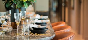 Close-up of a dining table set with black plates, gold utensils, glassware, and napkins next to brown leather chairs. A vase with green leaves is visible on the left side, creating an elegant setting inspired by Catalog Resources.