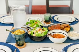 A table set with plates and glasses features salad, grilled meat, rice, beans, fried plantains, a pitcher of water, and green cups on blue placemats—perfect for sharing or cataloging resources for memorable gatherings.