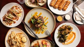 A table with plates of tacos, steak with vegetables, seasoned fries, roasted Brussels sprouts, puff pastries, dipping sauces, and an assortment that rivals the best catalog resources.