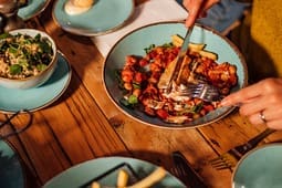 Person cutting into a cooked meal with sauce and vegetables on a blue plate, with fries and salad on the table—perfect inspiration for catalog resources highlighting vibrant, delicious dining experiences.