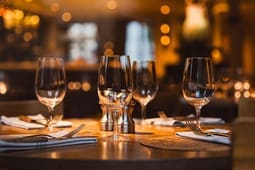 A set dining table with wine glasses, folded napkins, and cutlery in a softly lit restaurant evokes an atmosphere of elegance and refinement, making it an ideal scene for catalog resources.