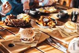 Plates with fried chicken, ribs, eggs, burgers, and drinks are arranged on a wooden table with people sitting around, ready to eat—like a feast inspired by the best catalog resources of comfort food favorites.