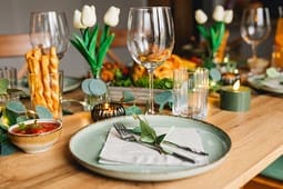 A dining table set with green plates, wine glasses, napkins, cutlery, candles, breadsticks, and white tulip decorations creates an inviting atmosphere inspired by Catalog Resources.