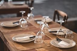 Four place settings on a wooden table with plates, napkins, utensils, upside-down wine glasses, and water glasses, ready for dining in an outdoor restaurant setting—perfect inspiration for Catalog Resources seeking stylish tablescape ideas.
