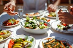 People sharing a meal outdoors, serving salad and various vegetable dishes from white plates on a sunlit table, while browsing catalog resources for fresh recipe inspiration.