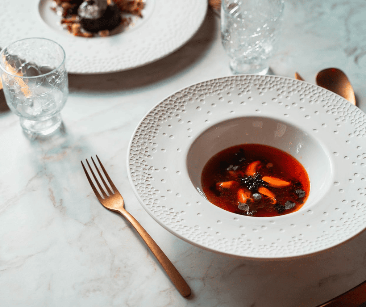 A white textured bowl of clear soup with carrots and garnish sits on a marble table, alongside a gold fork, knife, and two clear glasses.