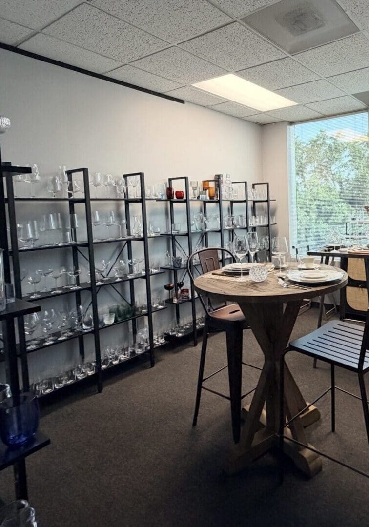 Room with shelves of glassware, a round wooden table set for two, and large windows letting in natural light, evoking the inviting elegance of a boutique showroom Houston is known for.