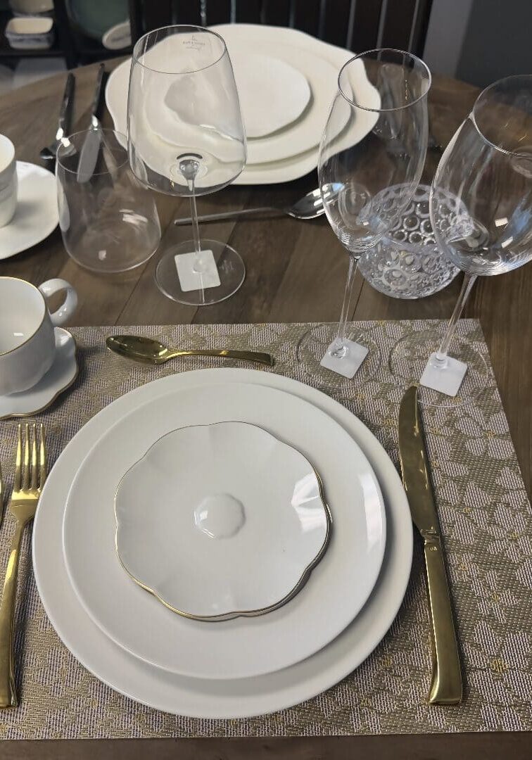 Formal table setting with white plates, gold utensils, wine glasses, and a teacup on a woven placemat atop a wooden table.